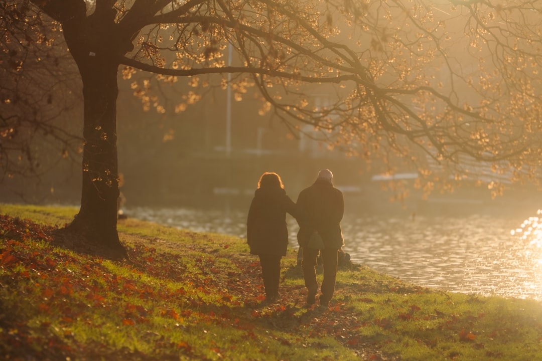 An older couple walks arm-in-arm along a grassy riverbank.