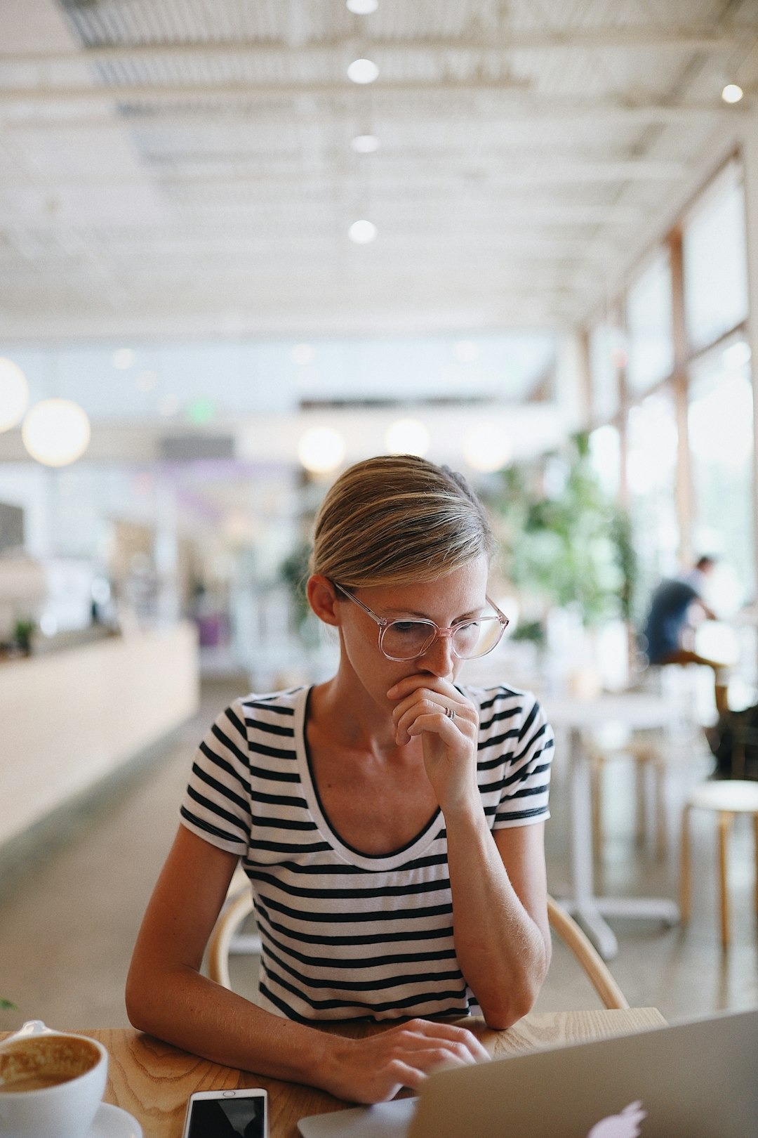 Woman in striped shirt thinking while working on laptop at cafe table.